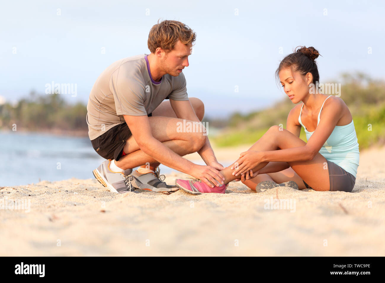 Injury - sports woman with twisted sprained ankle. Asian fitness female model sitting on beach sand getting help from Caucasian male touching her ankle. Stock Photo