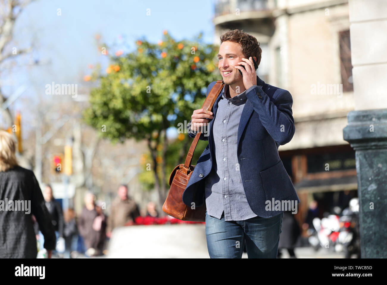 Young urban businessman on smart phone running in street talking on smartphone smiling wearing jacket and leather laptop bag on Passeig de Gracia, Barcelona, Catalonia, Spain. Stock Photo