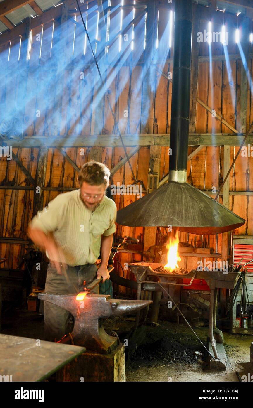 Light rays passing through wooden siding of blacksmith workshop, rural male blacksmith forge at work making metal shapes, Hillsboro, Wisconsin, USA. Stock Photo