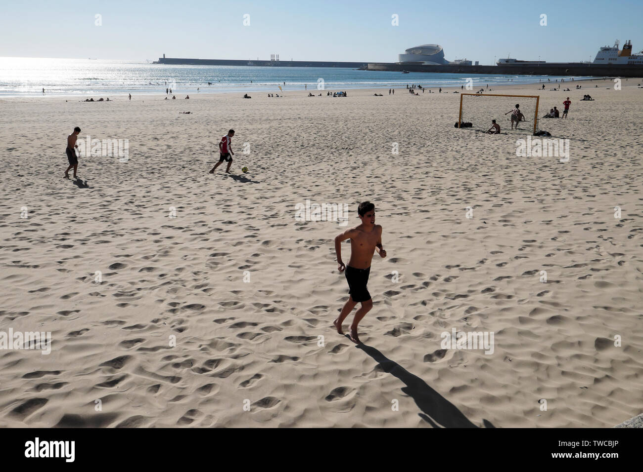 Young people playing football on beach in spring sunshine Matosinhos and view of Port of Leixões & Cruise terminal Porto Portugal Europe KATHY DEWITT Stock Photo