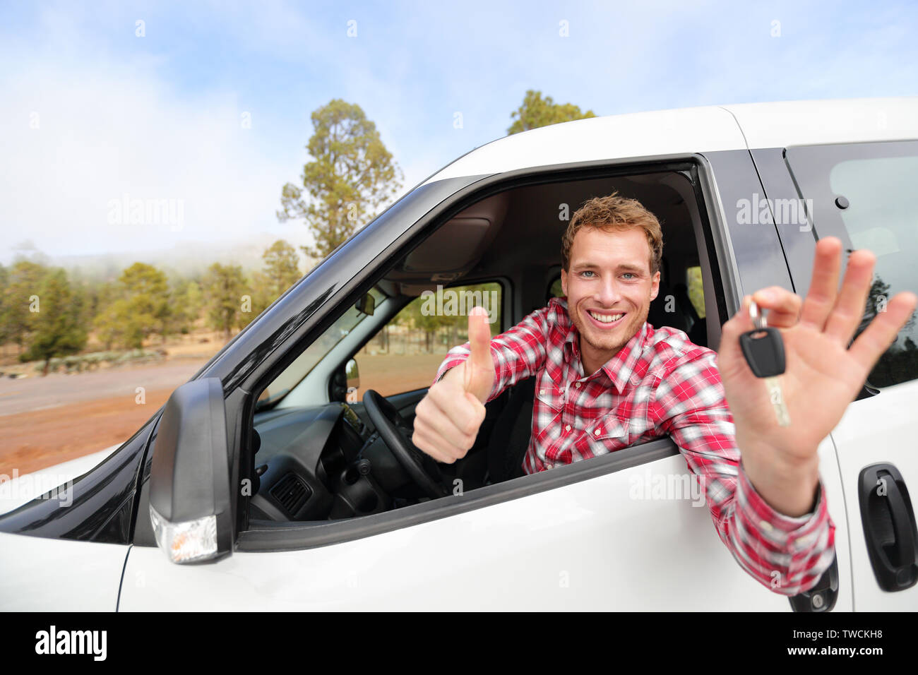 Car driver showing car keys and thumbs up happy. Young man holding car keys for new car. Rental cars or drivers licence concept with male driving in beautiful nature on road trip. Stock Photo
