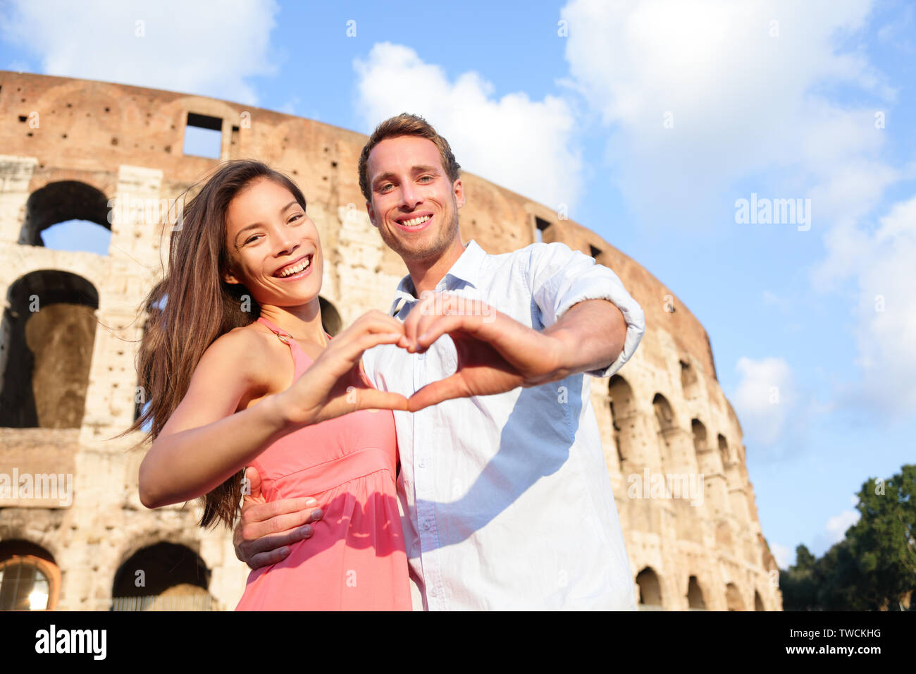 Romantic travel couple in Rome by Colosseum, Italy. Happy lovers on honeymoon showing heart sharped hands having fun in front of Coliseum. Love and travel concept with multiracial couple. Stock Photo