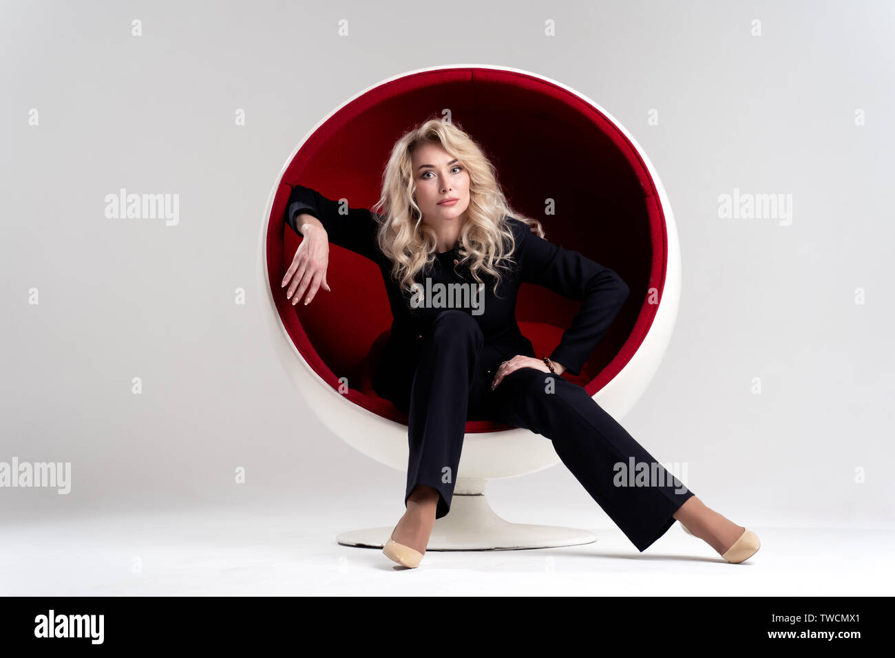Photo of blonde woman with long curly hair in black suit and beige shoes looking in camera while sitting in round chair isolated on white background Stock Photo