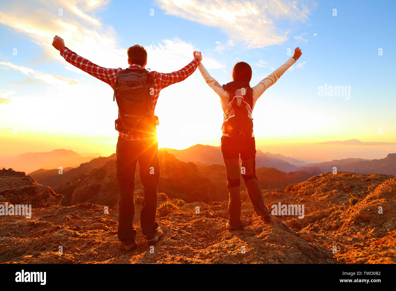 Freedom - Happy couple cheering and celebrating. Hiking man and woman raising arms excited in celebration outdoors. Hikers at sunset in mountain enjoying mountain top summit and success. Stock Photo