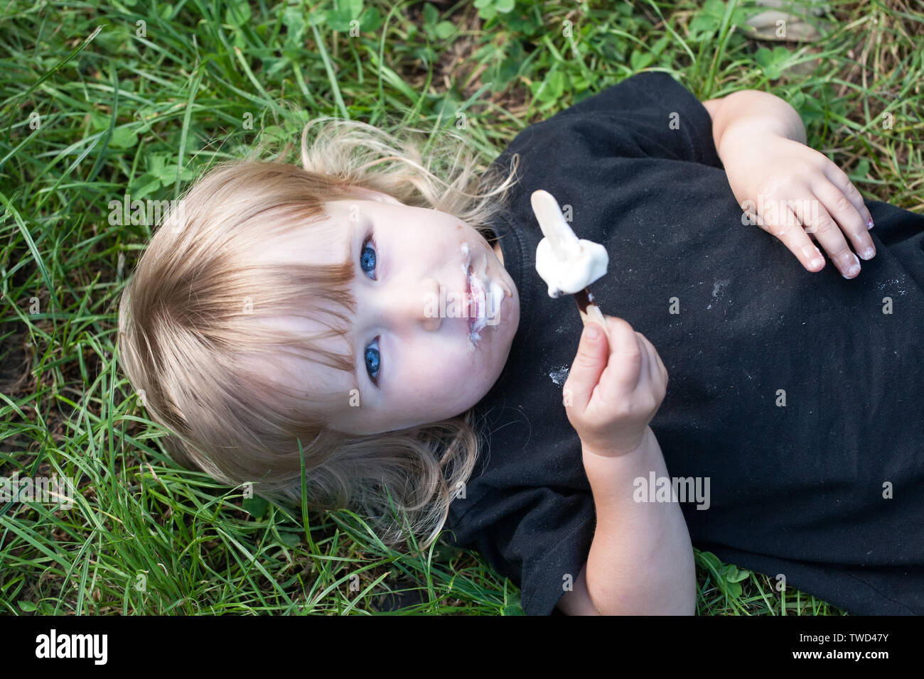 top view of little girl with chocolate popsicle ice cream in the hand lying on green grass closeup Stock Photo