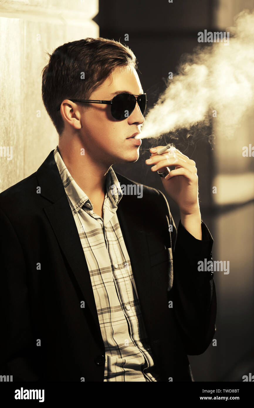 Young man in sunglasses smoking a cigarette on city street  Stylish fashion male model wearing black suit jacket Stock Photo
