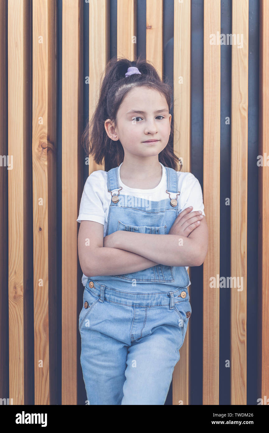 Cute happy caucasian girl in jeans overalls on a background of wall with wooden vertical slats. Lifestyle Stock Photo