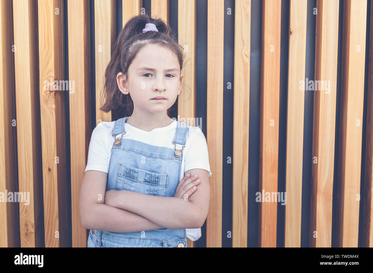 Cute happy caucasian girl in jeans overalls on a background of wall with wooden vertical slats. Lifestyle Stock Photo