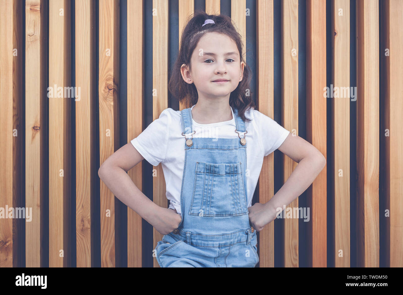 Cute happy caucasian girl in jeans overalls on a background of wall with wooden vertical slats. Lifestyle Stock Photo