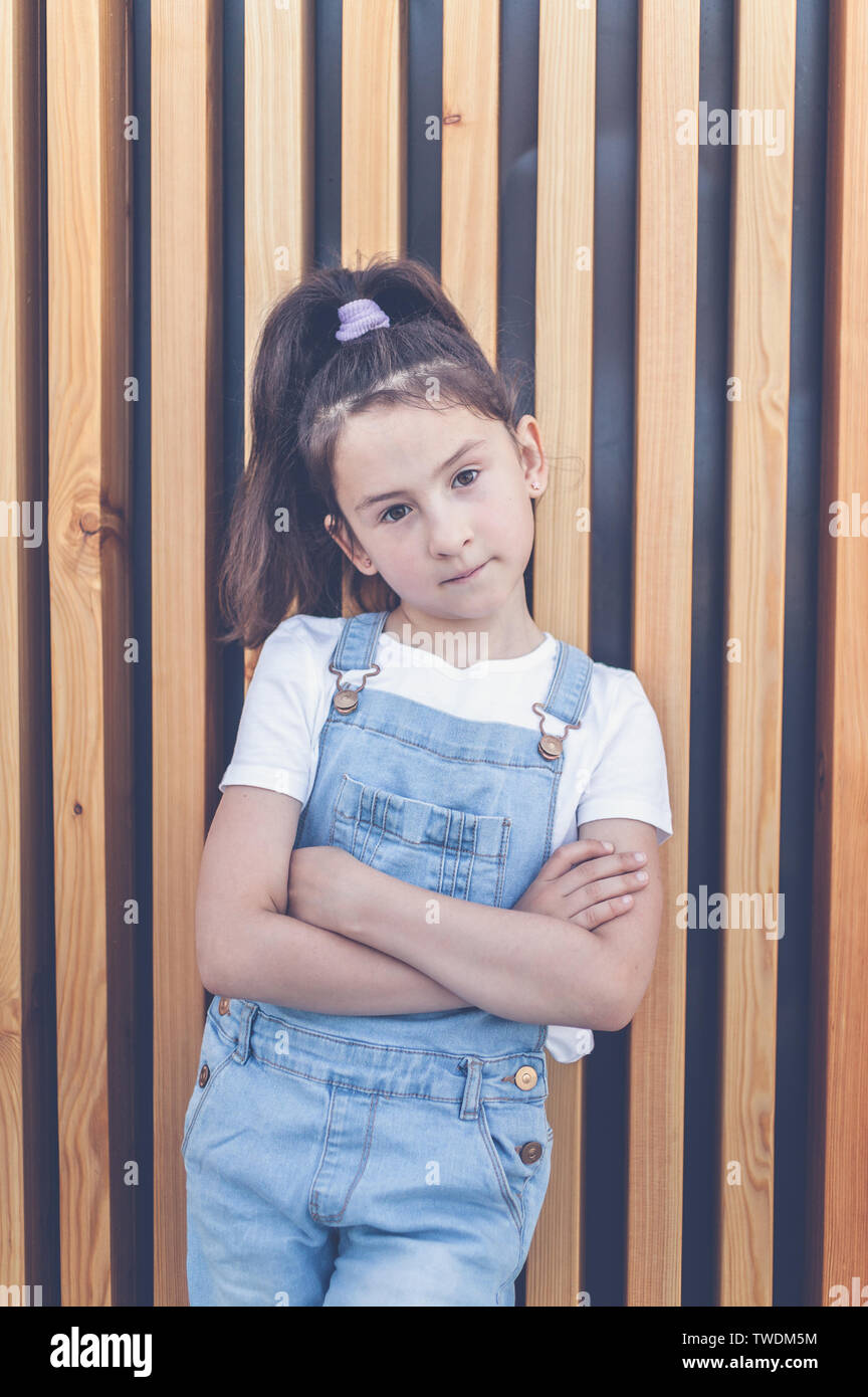 Cute happy caucasian girl in jeans overalls on a background of wall with wooden vertical slats. Lifestyle Stock Photo