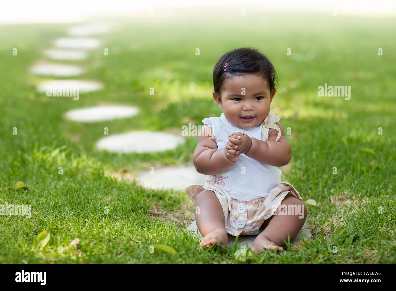 A baby girl grasping her own hands while sitting on the frontmost stepping stone from a series of stepping stones that go into perspective. Stock Photo