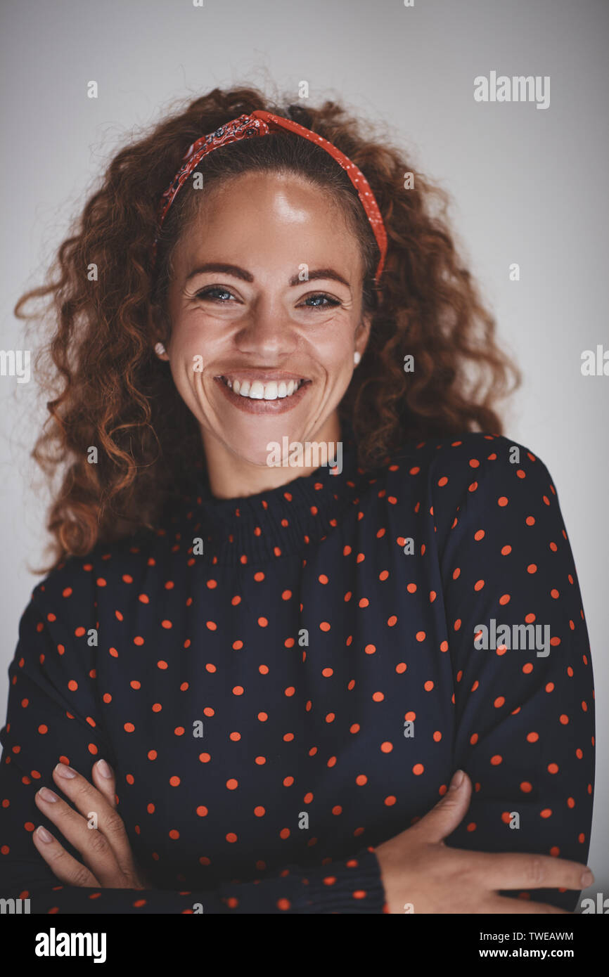 Casually dressed young female entrepreneur with curly hair smiling while standing with her arms crossed against a gray background Stock Photo