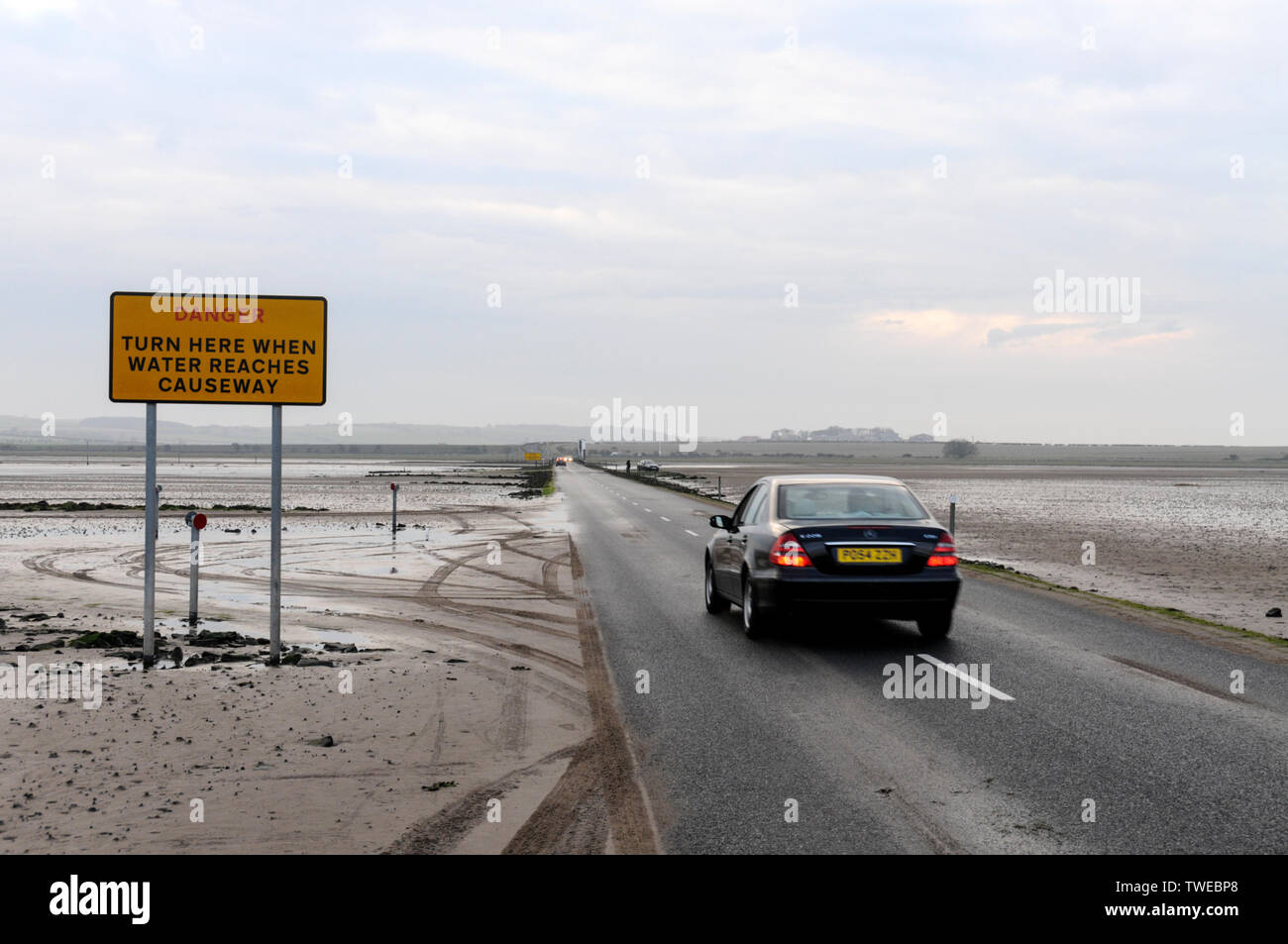 The Danger warning sign for tourist traffic crossing the 3-mile stretch of tarmac road at low tide connecting Lindisfarne Holy Island from the mainland Stock Photo