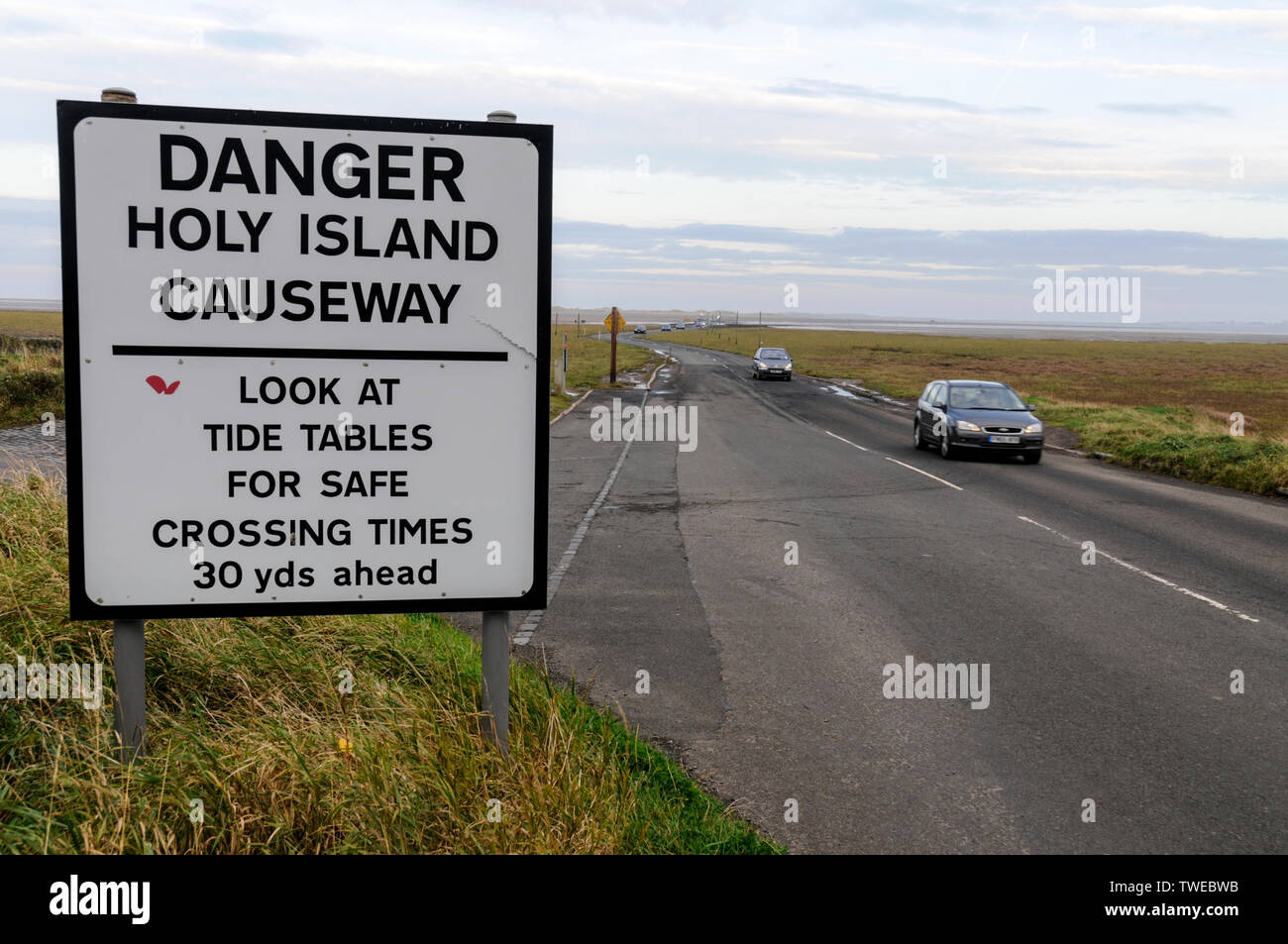 A road warning sign for visiting traffic crossing the 3 mile stretch of a narrow two way tarmac causeway at low tide connecting Lindistfarne Holy Isla Stock Photo