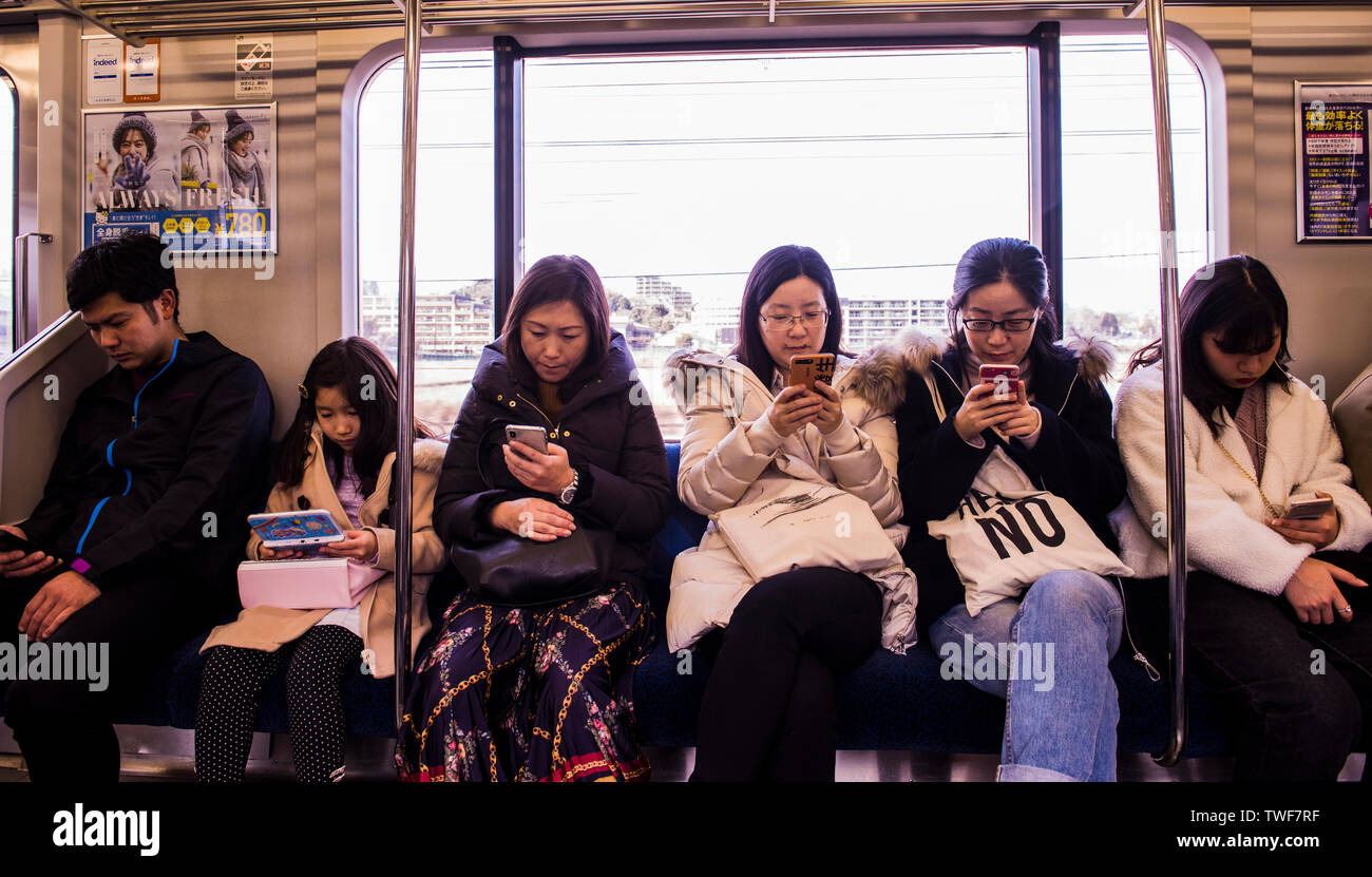 Row of people sitting on train on Tokyo underground with every person using either smartphone or electronic device. Stock Photo