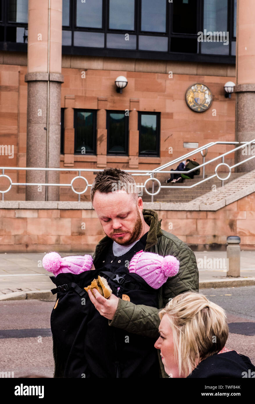 Couple outdoors with man carrying two young babies in body sling whilst eating sandwich in the Quayside area of Newcastle upon Tyne in England. Stock Photo