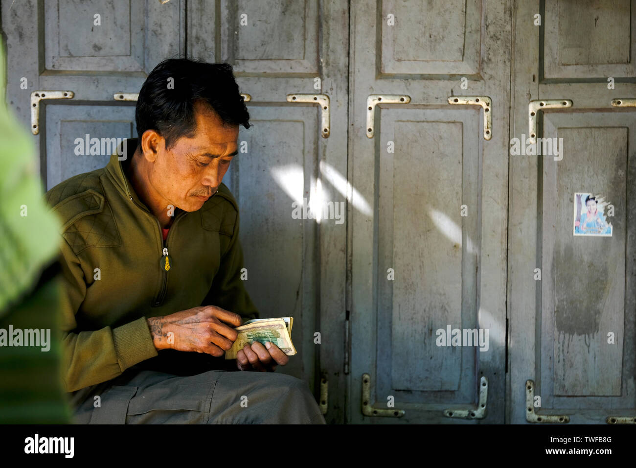 A Burmese market trader counts paper money in the shade of a tree. Stock Photo