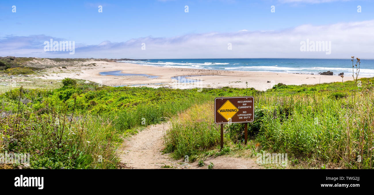 Walking path going through green shrubs towards a sandy beach; sign warning of Recurring Rip Currents posted on the trail; Gazos Creek Año Nuevo State Stock Photo