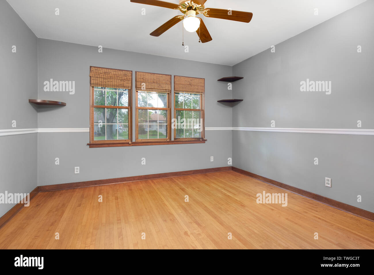 A plain grey room with with hardwood floors, ceiling fan and a view to a residential neighborhood. Stock Photo