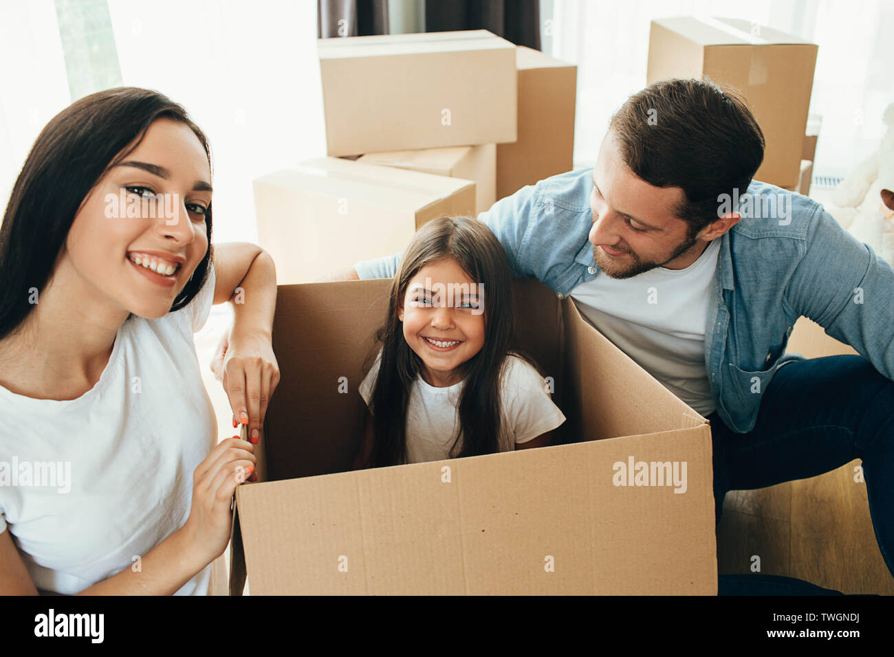 Happy family having fun while moving into new apartment. Mother, father and daughter together smiling, with Cardboard boxes on background Stock Photo