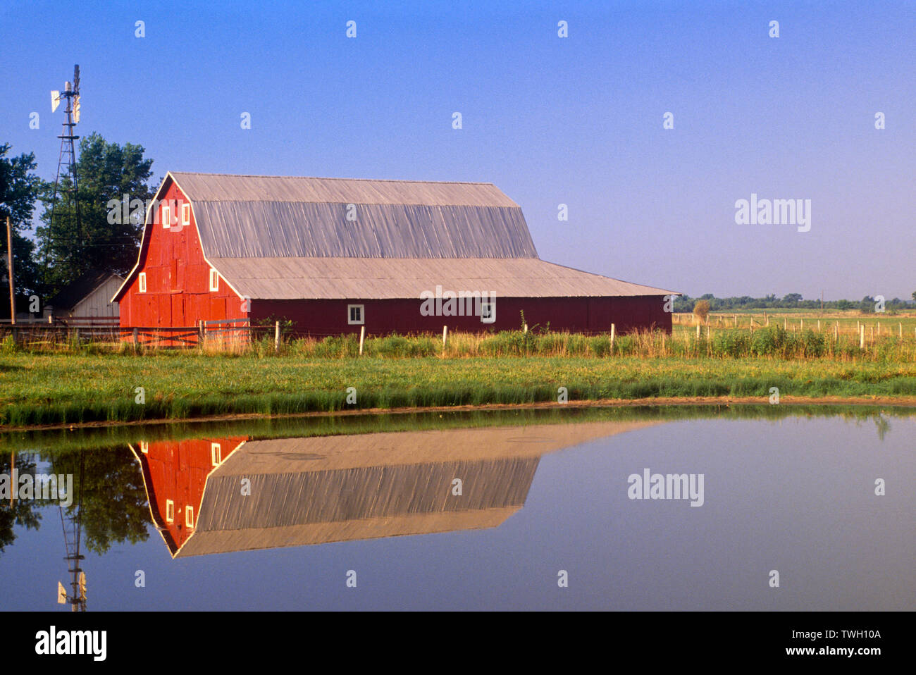 A classic red barn with windmill reflected in pond in a rural midwest on a blue sky summer day, Missouri, USA Stock Photo
