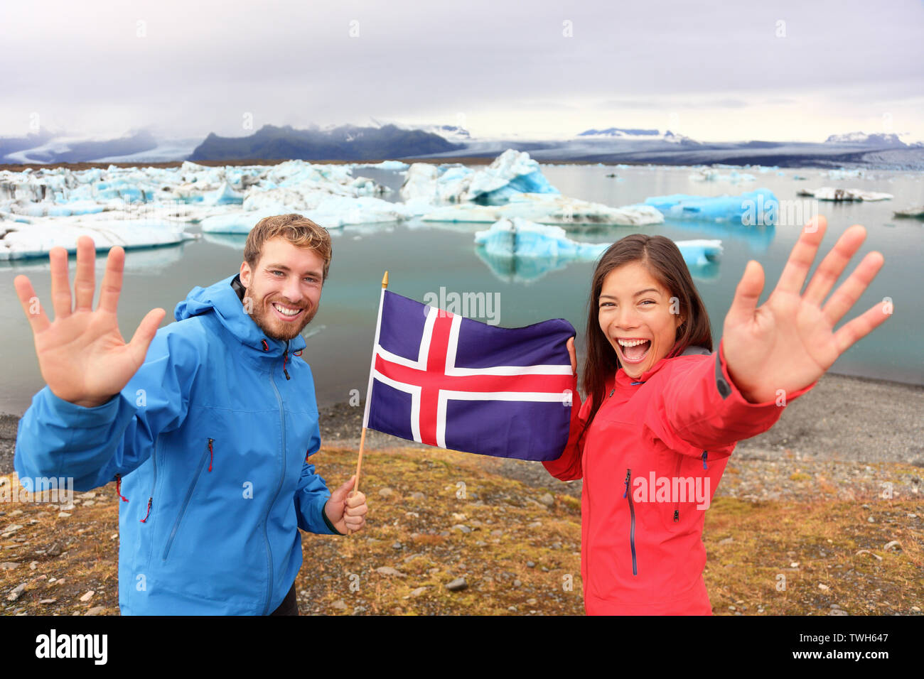 Icelandic flag - tourists on Jokulsarlon, Iceland on travel. Tourist couple happy holding showing Icelandic flag in front of the glacial lake / glacier lagoon. Stock Photo