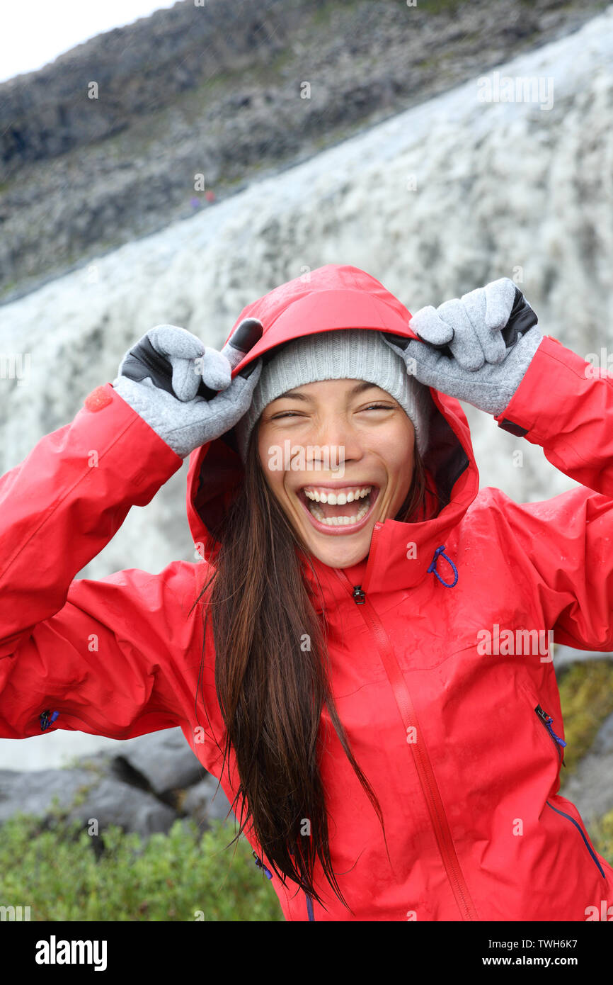 Woman laughing in raincoat by Dettifoss waterfall on Iceland. Excited girl in hardshell visiting tourist attractions and landmarks on Diamond Circle. Dettifoss waterfall in Vatnajokull National Park. Stock Photo
