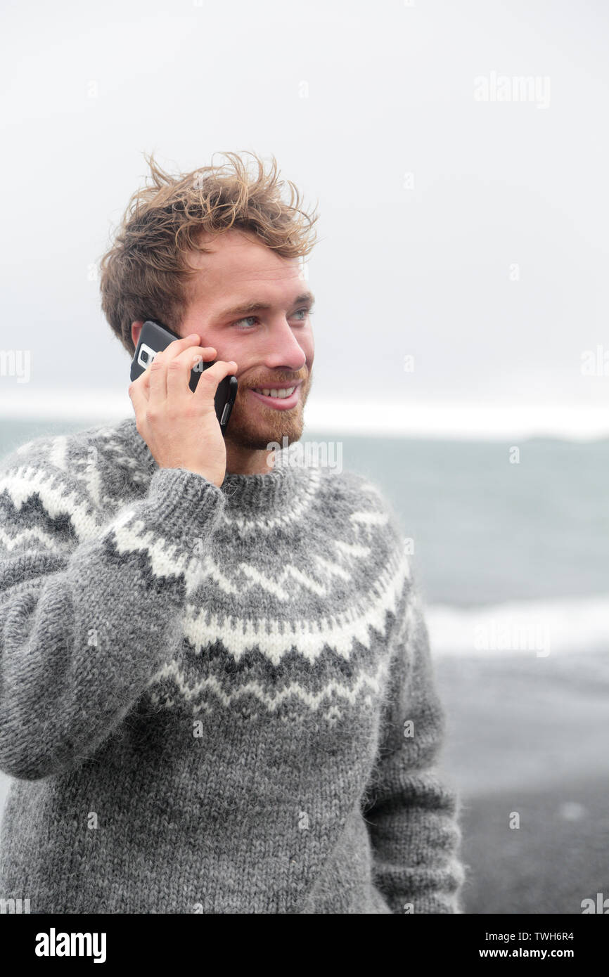 Smartphone man talking on smart phone walking on black sand beach on Iceland wearing Icelandic sweater by the ocean sea. Stock Photo
