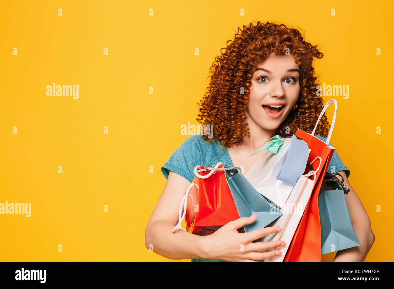 Curly haired woman with surprised face holding many shopping bags on yellow background Stock Photo