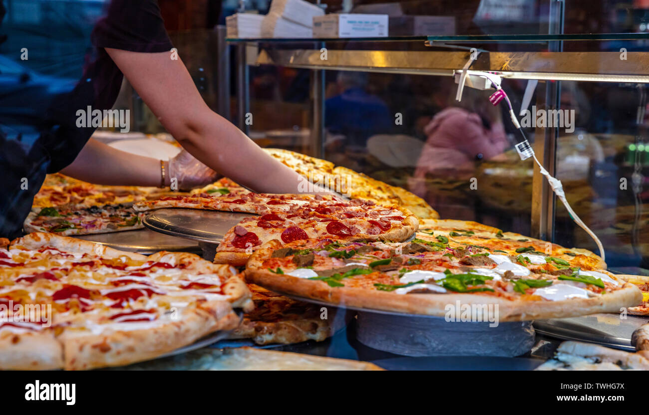 USA, New York. May 3, 2019. Pizzeria glass window. Variety of italian pizzas in a take away shop display, street food. Kitchen female worker hands arr Stock Photo