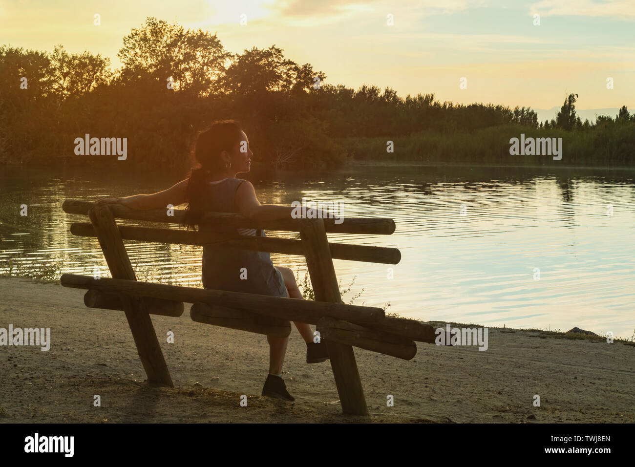Young brunette woman watching the sunset, sitting on a wooden bench in front of a Tajo river, Spain Stock Photo