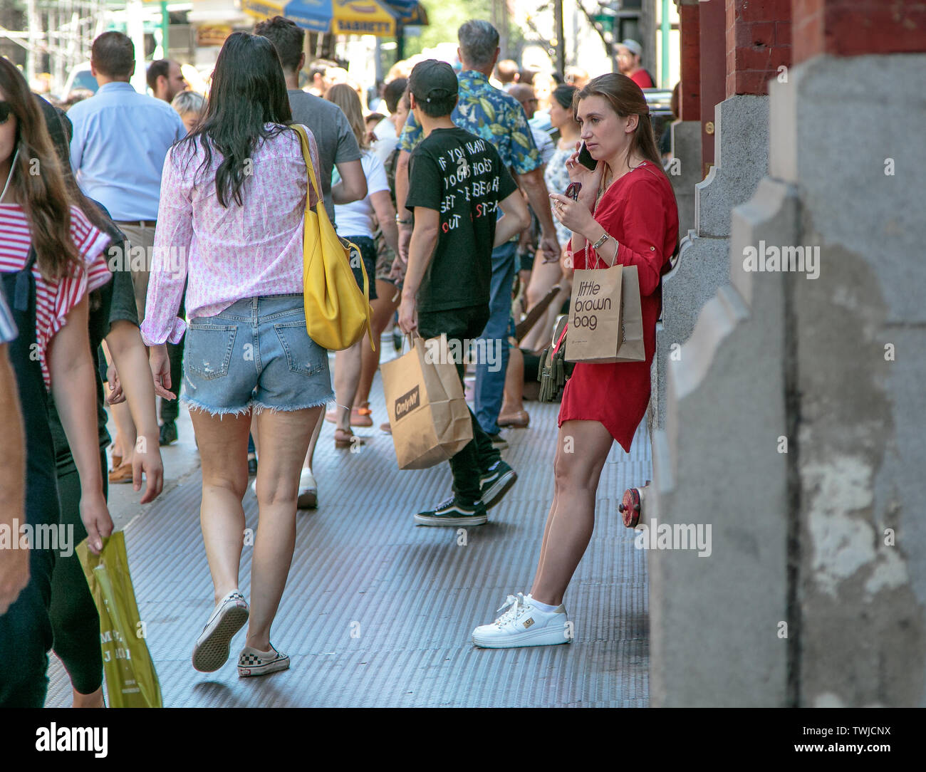 New York, 6/15/2019: Unidentified young lady in a red dress taks on the phone while leaning against a building wall and holding a shopping bag from Bl Stock Photo