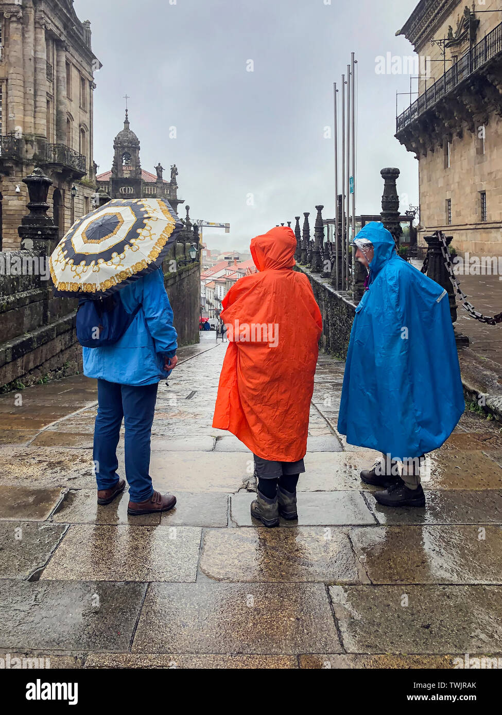 Three pilgrims back to us, with backpacks in colorful raincoats with an umbrellas standing in the historic square Stock Photo