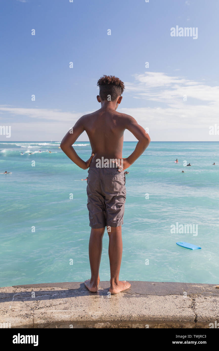 A scene from the beach.  On sunny day, a young boy standing on a ledge looking down water below at Waikiki beach in Hawaii. Stock Photo