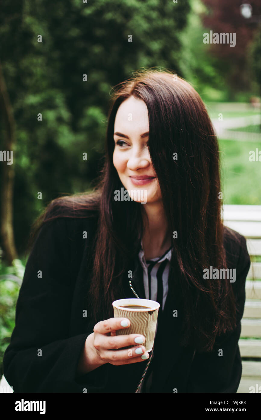 Cute smiling brunette holding a paper cup of black coffee and looking in distance. Charming business woman wearing a black coat and striped black and Stock Photo