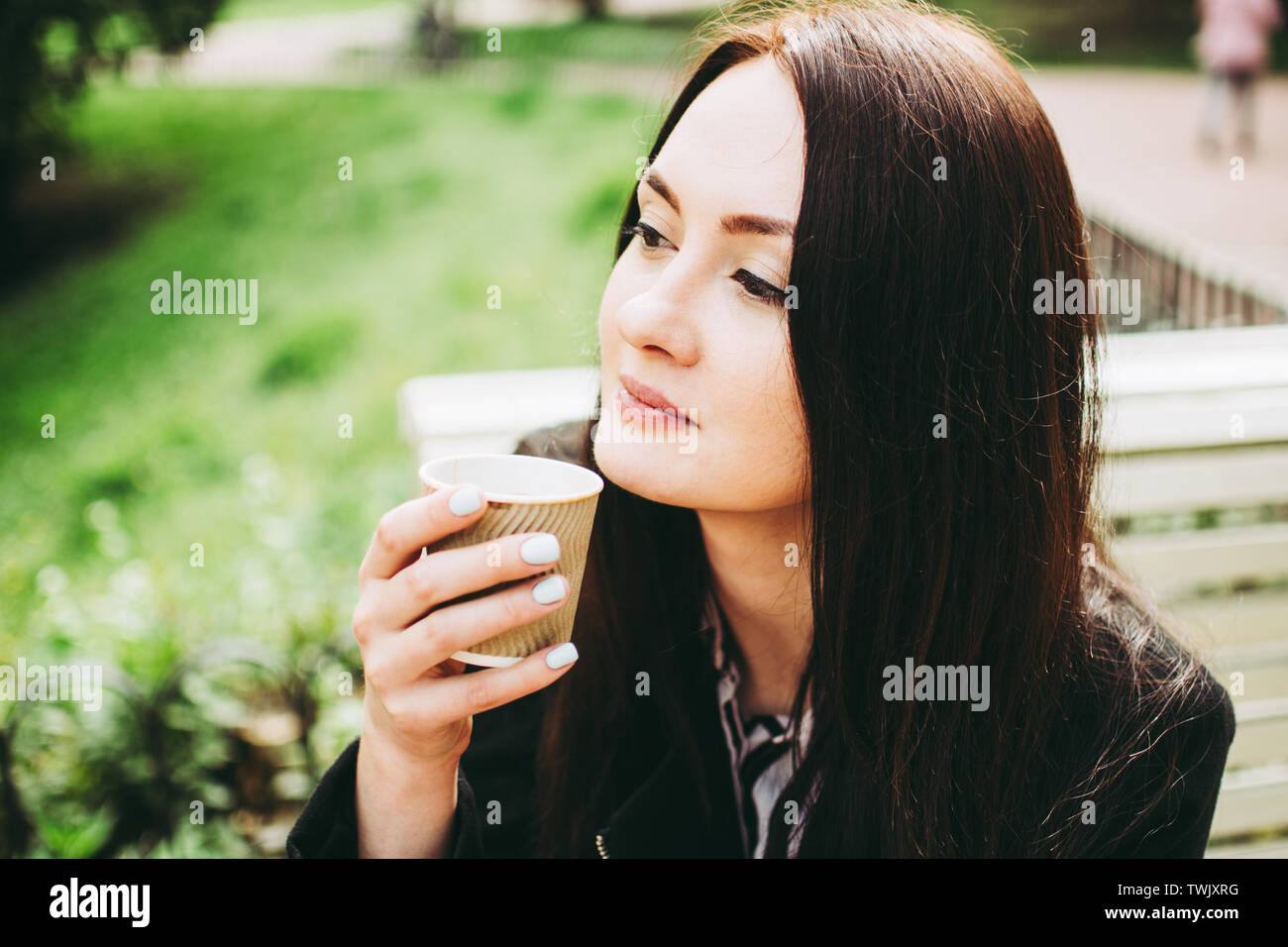 Cute smiling brunette holding a cup of coffee and smelling the aroma. Charming business woman sitting on a bench in a park. Stock Photo
