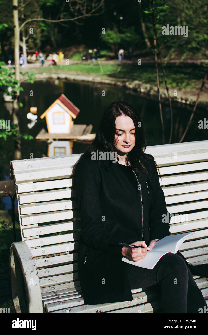 Pale skinned brunette working on her homework assignment (essay or research paper) and writing down her thoughts in a notebook. Lake and wooden house Stock Photo