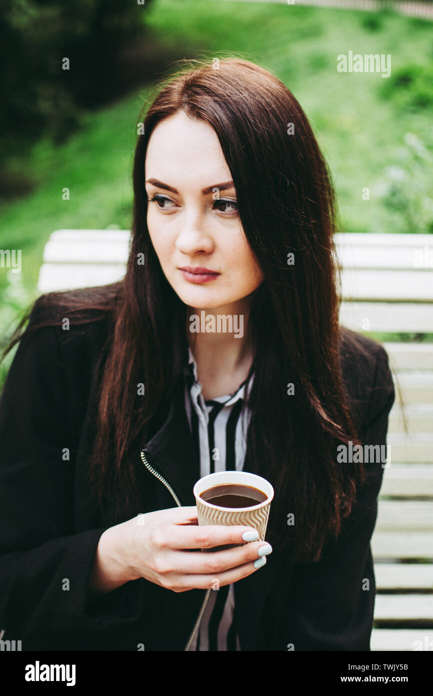 Gorgeous black-eyed brunette holding a paper cup of black coffee and looking in distance. Charming business woman wearing a black coat and striped bla Stock Photo