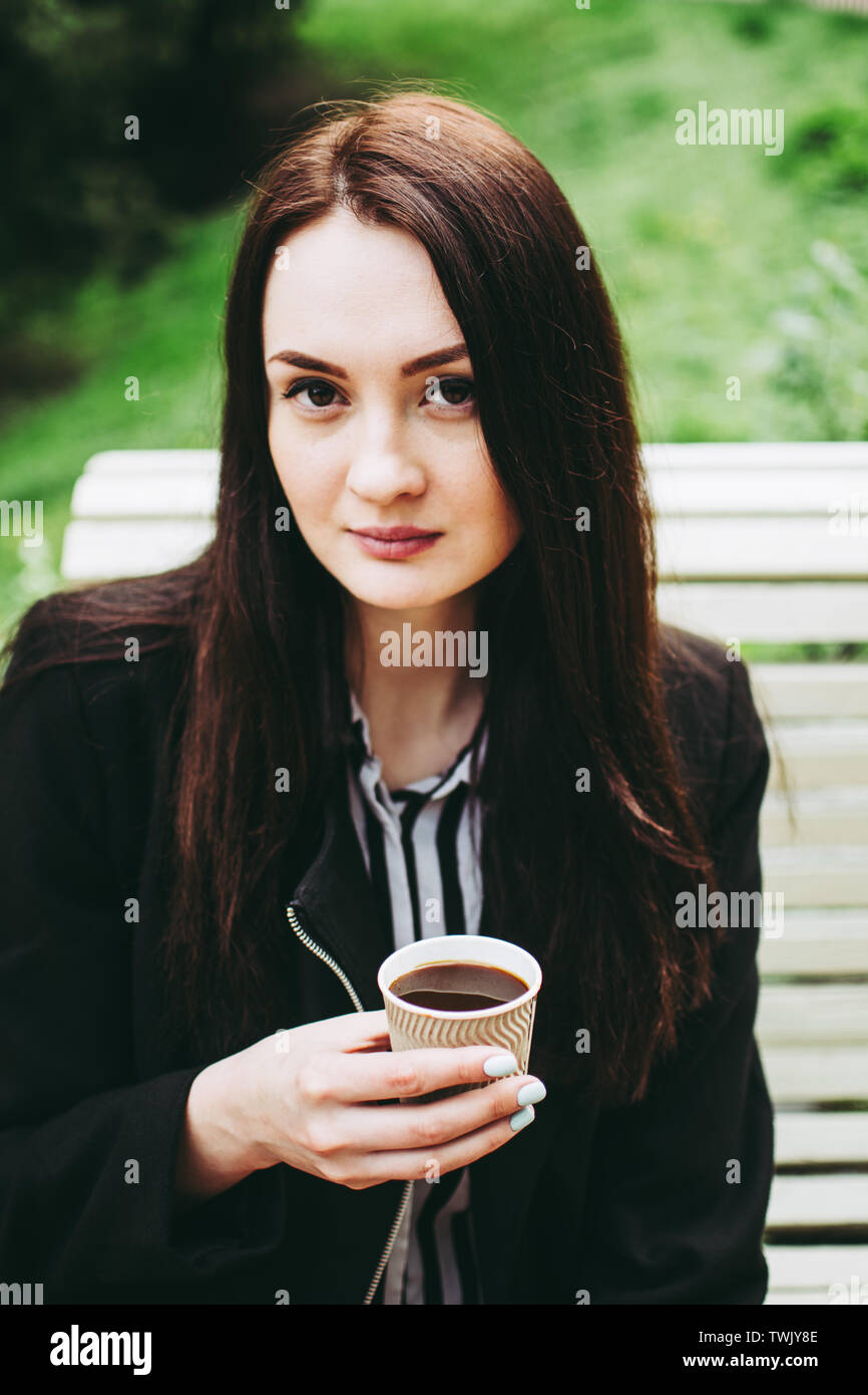 Gorgeous black-eyed business woman holding a paper cup of black coffee and looking at the camera. Charming young brunette wearing a black coat and str Stock Photo
