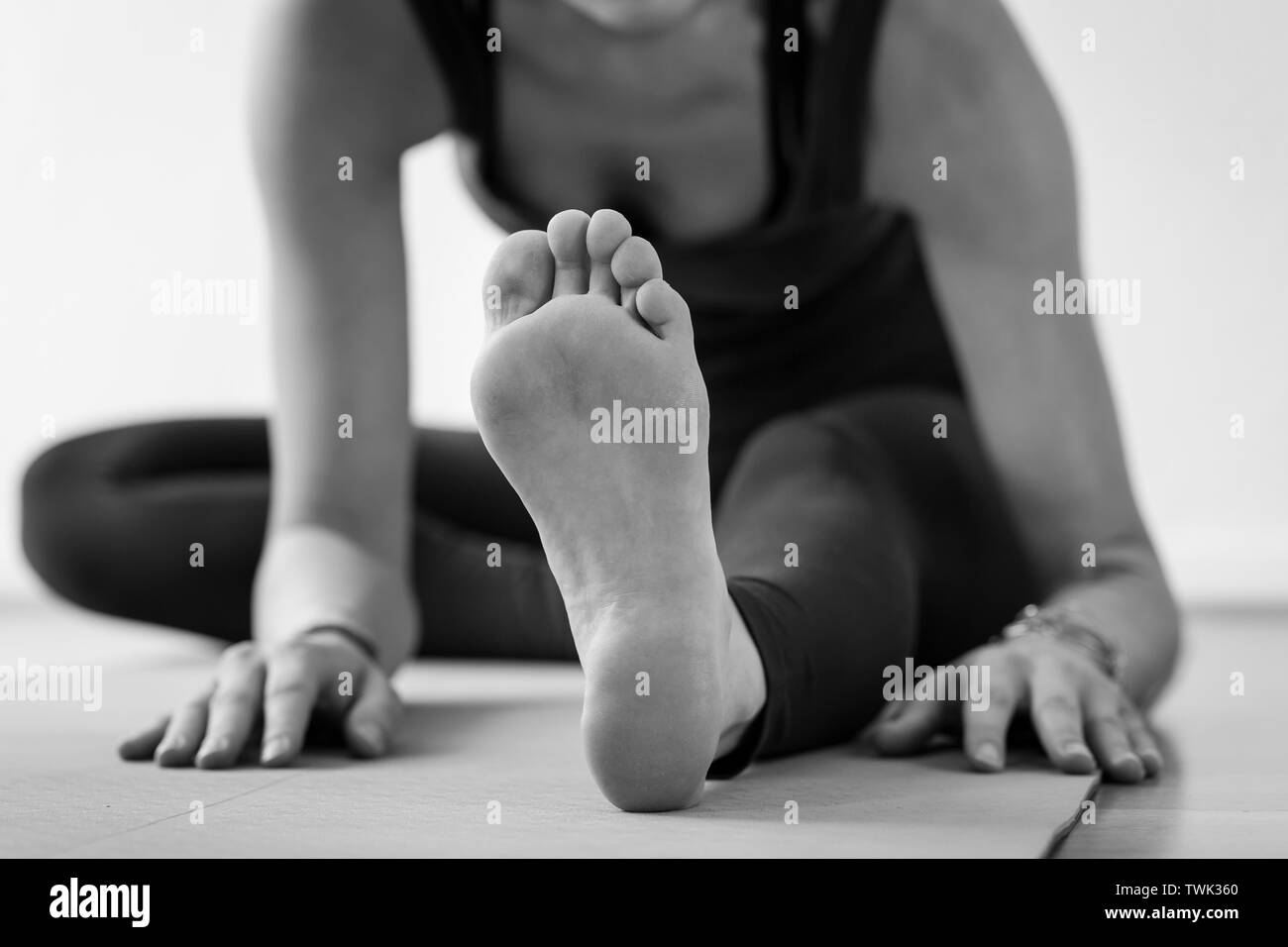 Unrecognizable European woman on the floor on her way to Janu Sirsasana or Head-to-Knee Forward Bend, aka seated yoga pose. Black and white horizontal Stock Photo