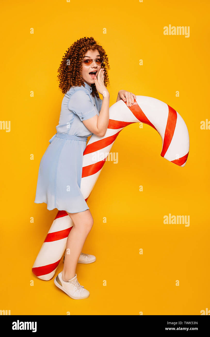 surprised curly-haired woman shocked covering her mouth standing with sweet big candy cane on yellow background Stock Photo