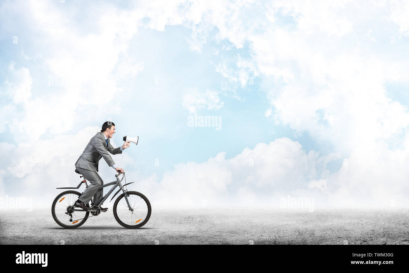 Businessman with megaphone on bike at sunny day. Marketing and advertising campaign. Manager in business suit riding bicycle and speaking in loudspeak Stock Photo