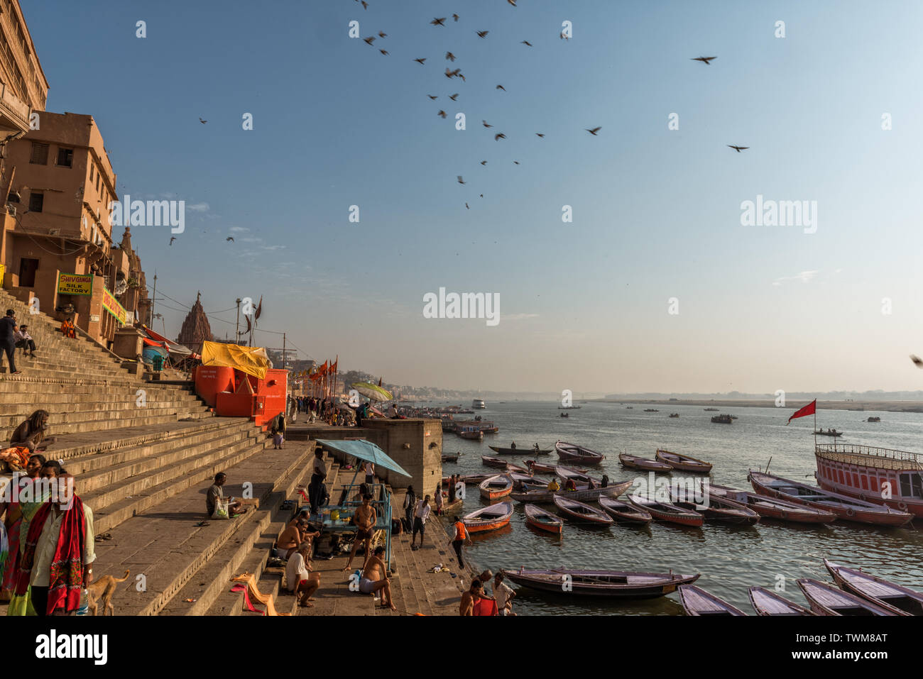 Varanasi Ghat on a winter soft sunny morning with boats and the pilgrims moving around the ghats. Morning birds are flying in deep blue skyof Varanasi Stock Photo