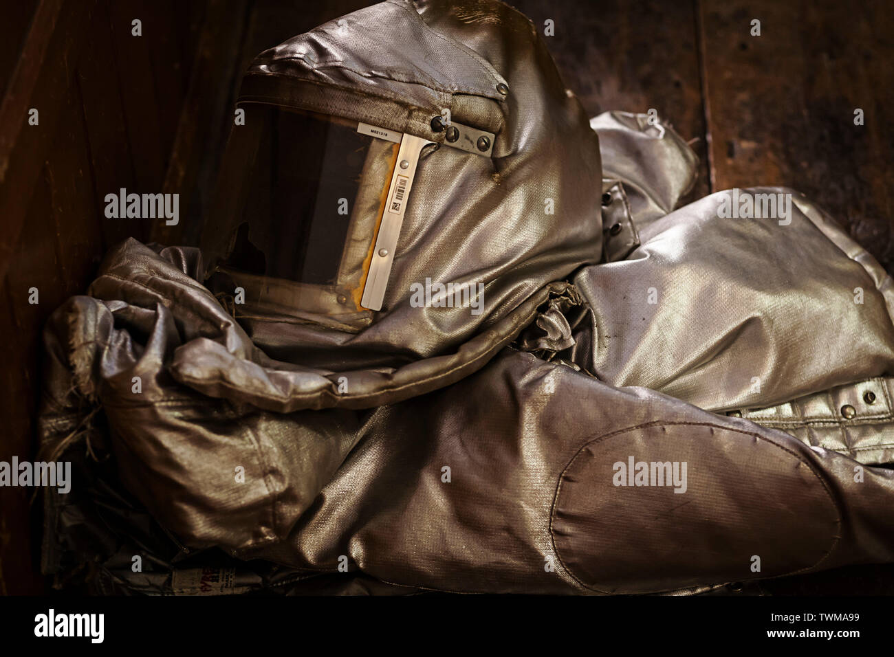 A folded up slightly damaged fire proximity suit also known as a bunker suit on a wooden floor. There is a visible hole in the visor of the suit. Stock Photo