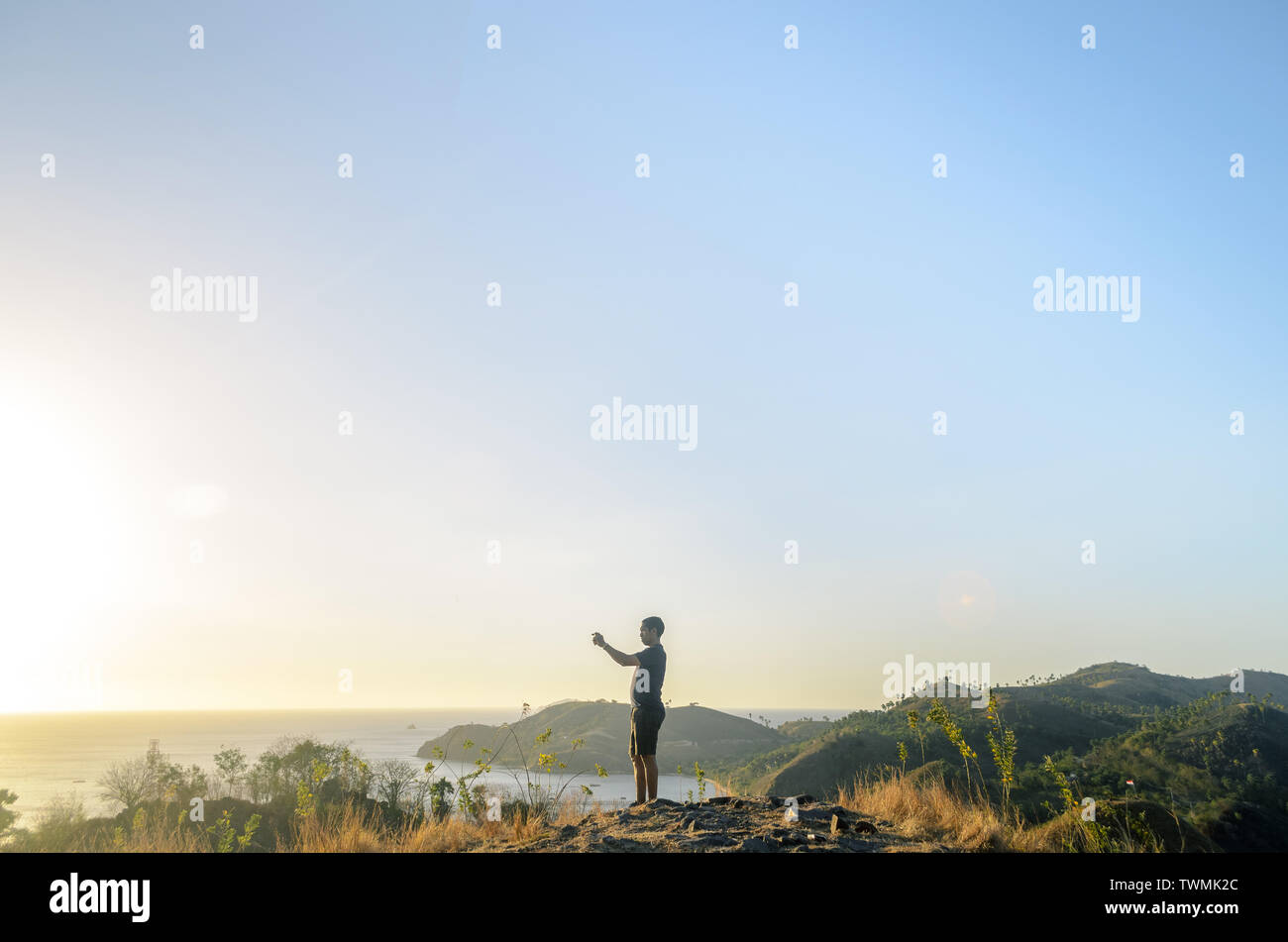 a tourist taking photograph of sunset with a smartphone from a hill with sea and blue sky in the background, at Labuan Bajo, Indonesia Stock Photo