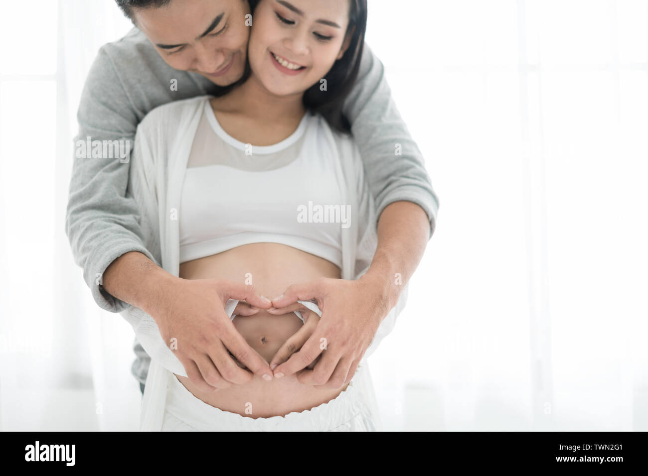 Asian beautiful pregnant woman and her handsome husband hugging the tummy. Love couple hands make a heart on the pregnant belly. Stock Photo