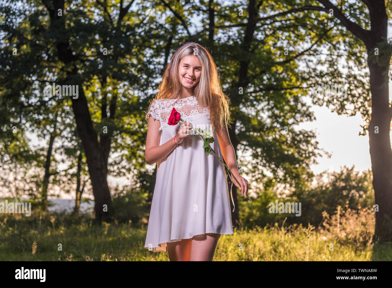 The smiling model in front of the camera. Innocent, with a rose in her hand (a symbol of love), with a beautiful backdrop, with blonde hair gently tou Stock Photo