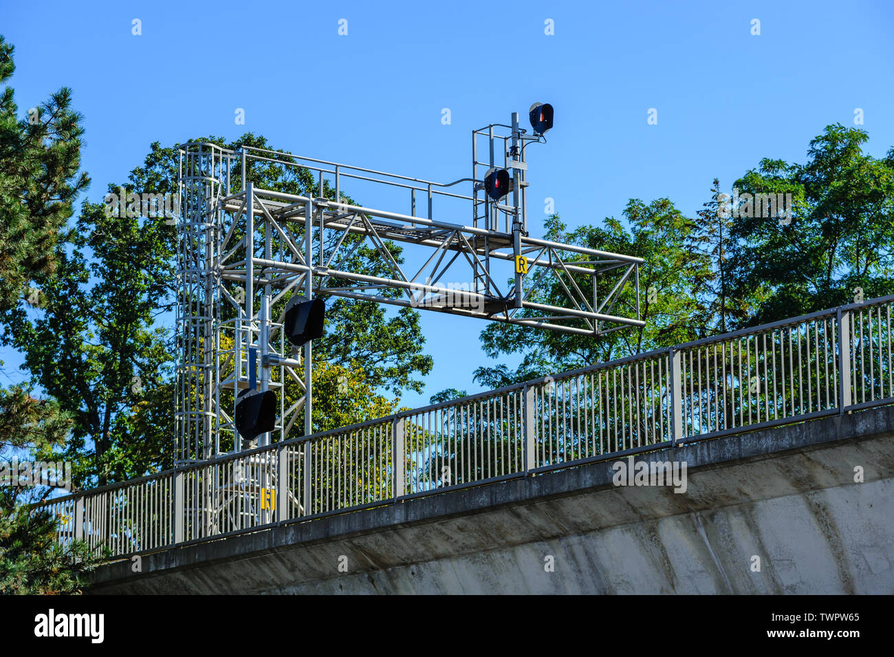 Railway signals and signs on metal frame among trees above bridge with fence. Stock Photo