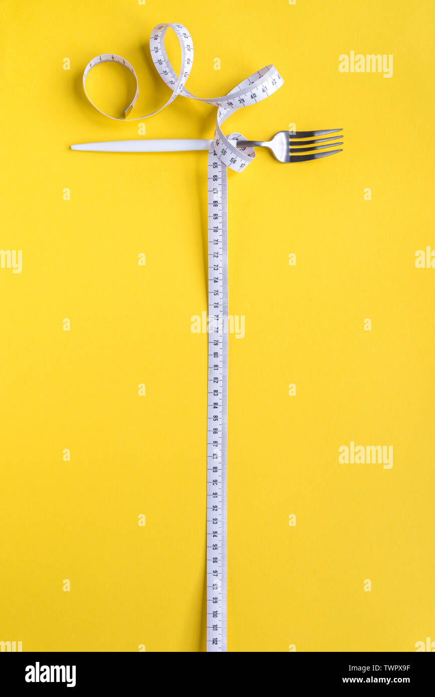 Fork and Measuring Tape on yellow background, copy space. Diet, healthy lifestyle, weight loss concept with white fork and measuring tape. Stock Photo