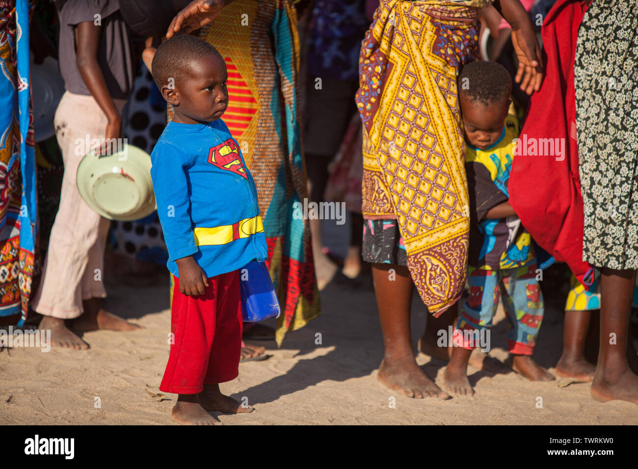 A hungry child wearing a Superman suit waits in line to get food on a hot day with other children in the background Stock Photo